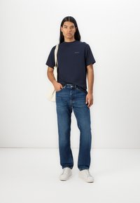 Marineblå crewneck t-skjorte kombinert med blå rette jeans og hvite joggesko. Personen holder en lys beige tote bag.