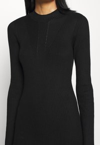 Schwarzes Rippstrickkleid mit hohem Ausschnitt, langen Ärmeln und vertikalen Nahtdetails am Oberteil. Strukturierter Stoff verleiht Tiefe.