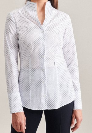 Vrouw draagt een nauwsluitend wit overhemd met kleine blauwe stippen en een kleine geborduurde bloem bij de taille, gecombineerd met een zwarte broek.