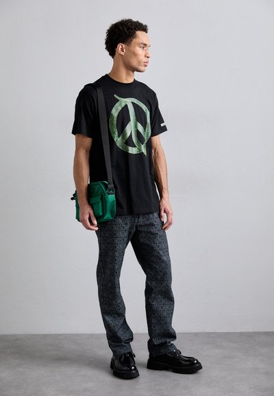 T-shirt noir avec un grand graphique de symbole de la paix vert, associé à un pantalon de survêtement noir à motifs et un sac à bandoulière vert. Chaussures noires.