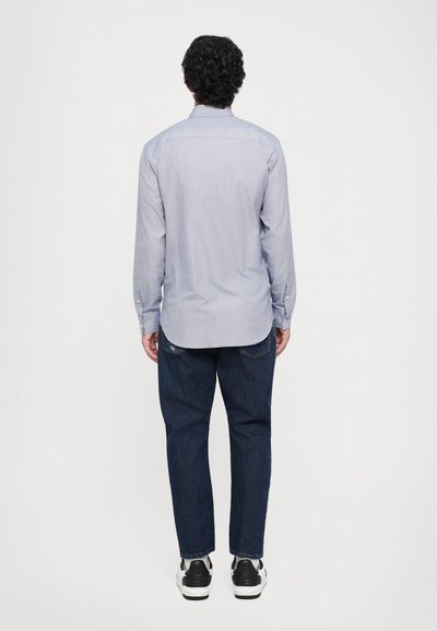 Mann mit lockigem, dunklem Haar steht mit dem Rücken zur Kamera, trägt ein hellblaues Langarmshirt, dunkle Jeans und weiße Sneaker mit schwarzen Akzenten.