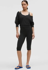 Černá oversized mikina, zkrácená v pase, spárovaná s černými legínami. Modelka má na sobě modré žabky. Hladká látka s bílými akcenty.