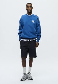 Blå sweatshirt med vit logga, kombinerad med mörka shorts. Modellen bär vit skjorta med krage under, vita strumpor och grå sneakers.