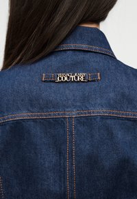 Σκούρο μπλε τζιν μπουφάν με πορτοκαλί ραφές που φοριέται από ένα άτομο με μακριά σκούρα μαλλιά, με χρυσή ετικέτα «Versace Jeans Couture» στην πλάτη.