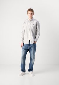 En ung mann står iført en lys grå skjorte med knapper, blå jeans og hvite sneakers foran en enkel hvit bakgrunn.