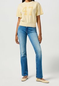 Frau trägt ein hellgelbes, kurzes T-Shirt mit der Nummer 47, ausgewaschene, gerade blaue Jeans und beige Ballerinas, während sie vor einem schlichten Hintergrund steht.