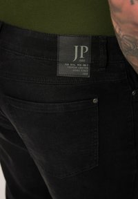 Jeans de mezclilla negra con una superficie texturizada, que muestran un bolsillo trasero cosido y una etiqueta rectangular con "JP 1880" en letras verdes.