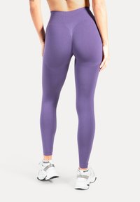 Vista trasera de una persona que lleva leggins morados de talle alto y zapatillas blancas, de pie con las manos relajadas a los lados sobre un fondo blanco.