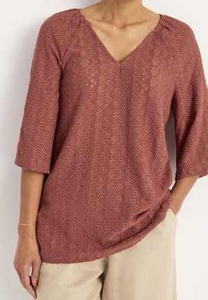 Vrouw draagt een stoffige roze, getextureerde V-hals blouse met driekwart mouwen en lichtbeige broek, één hand in de zak.
