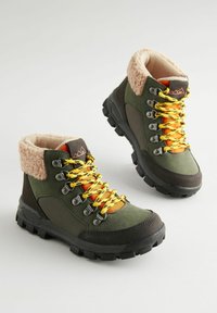 Zielone buty trekkingowe z brązowymi akcentami, kołnierzem z sztucznego futra, żółtymi i czarnymi sznurowadłami oraz gumową podeszwą, z metalowymi oczkami dla wsparcia sznurowania.