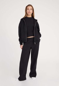 Schwarze Zip-Hoodie mit Kapuze, langen Ärmeln und zwei vorderen Taschen; kombiniert mit locker sitzenden schwarzen Jogginghosen. Glatte Stoffstruktur.