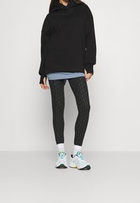 Svart oversized hoodie, lager över en blå topp, kombinerad med mönstrade svarta leggings och vita sneakers med färgglada accenter.