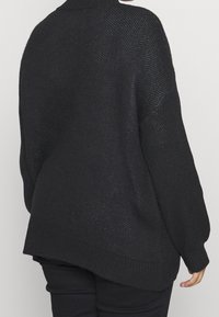 Pull en tricot noir avec une coupe ample, des épaules tombantes et des poignets côtelés. Présente un motif en tricot ouvert et un ourlet arrière plus long.
