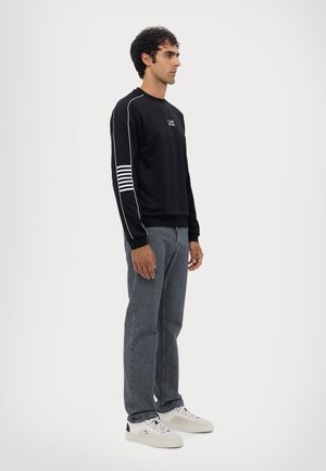 Felpa nera realizzata in materiale morbido con strisce bianche sulla manica sinistra e un logo sul petto, abbinata a jeans scuri e sneaker bianche.