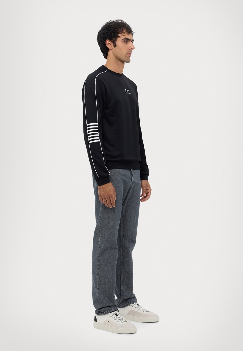 Felpa nera realizzata in materiale morbido con strisce bianche sulla manica sinistra e un logo sul petto, abbinata a jeans scuri e sneaker bianche.