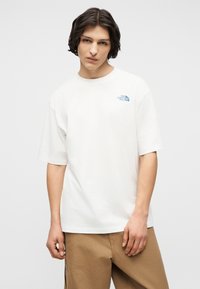 Νεαρός άνδρας με μεσαίου μήκους σκούρα μαλλιά που φοράει λευκό μπλουζάκι The North Face και ανοιχτό καφέ παντελόνι, μπροστά σε απλό φόντο.