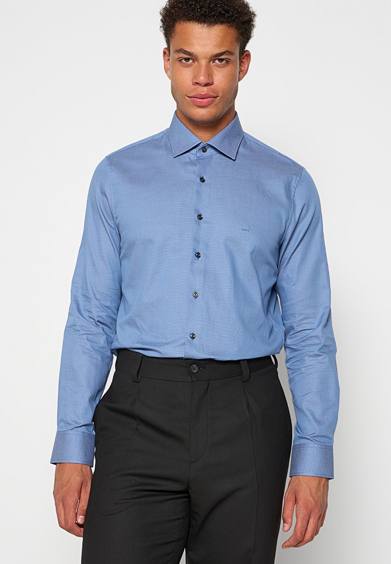 Chemise de couleur bleu clair avec un col boutonné, des boutons noirs et un tissu texturé. Portée avec un pantalon de costume noir.