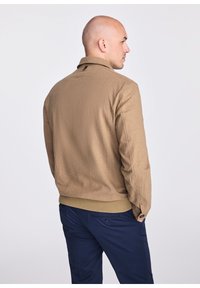 Brązowa kurtka bomberka z teksturowanej tkaniny, z prążkowanymi mankietami i dołem. Posiada kołnierz i mały akcent w postaci logo w okolicy szyi.