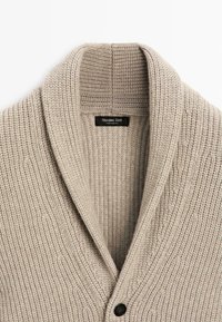 Rippstrick-Cardigan in hellem Beige mit breitem Schalkragen und einem schwarzen Knopf vorne. Etikett sichtbar im Kragen.