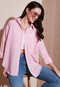 Lightweight, pink and white striped button-up shirt with a pocket, featuring rolled-up sleeves and a relaxed fit. Worn over a white top.