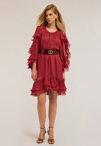 Žena v burgundských šatech s volány a průhlednými vrstvenými rukávy, s tmavým širokým páskem a hnědými sandály na vysokém podpatku na jednobarevném pozadí.