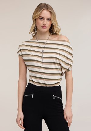 Blonde Frau, die ein schulterfreies, beige und gold gestreiftes Oberteil, schwarze Hosen mit Reißverschlusstaschen, eine lange Anhänger-Halskette und hängende Ohrringe trägt.