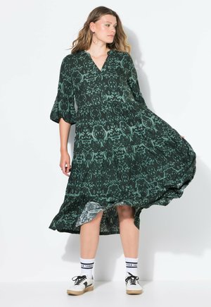 Robe verte à motifs avec un décolleté en V et des manches évasées, fabriquée en tissu léger, avec un design à volants et un ourlet évasé. Portée avec des baskets.