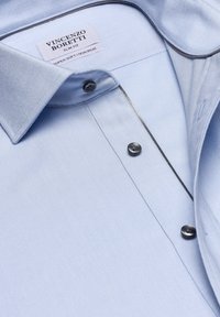 Helles blaues Slim-Fit-Hemd mit glatter Textur, schwarzen Knöpfen, kontrastierendem Besatz am Kragen und einem "pflegeleicht"-Label ohne Bügeln.
