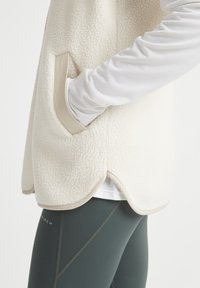 Fleecejacka i off-white med en texturerad yta, med en beige fickdetalj och rundad nederkant, kombinerad med mörkgröna leggings.