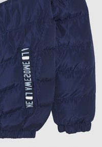 Giacca puffer navy con texture trapuntata, polsini elasticizzati e un accento del logo in bianco sul lato.