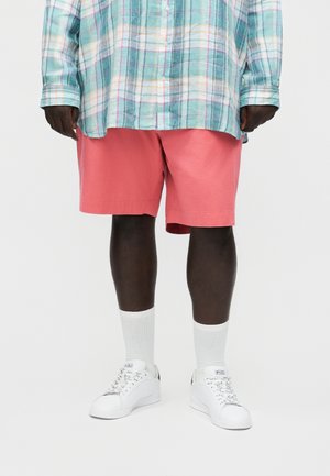 Άτομο που φοράει κοραλί ροζ σορτς, λευκές κάλτσες, λευκά αθλητικά παπούτσια Polo και μία φαρδιά, πολύχρωμη καρό πουκάμισο με κουμπιά, στέκεται μπροστά σε λευκό φόντο.