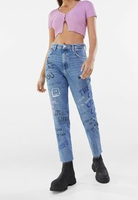 Top corto de canalé rosa claro con mangas cortas combinado con unos vaqueros de cintura alta que presentan varios gráficos de texto en negro y de colores. Botas negras hasta el tobillo.