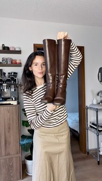 Bottes en cuir marron montant jusqu'aux genoux, maintenues en l'air, présentant une texture lisse, un bout arrondi et une fermeture éclair latérale. Exposées contre un intérieur neutre.
