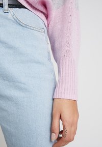 Manche de pull tricoté en tons roses et gris, poignets côtelés, associée à un jean denim bleu clair. Main visible, affichant un petit tatouage.