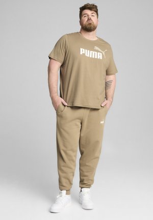 Tan PUMA t-shirt with logo across the chest, paired with matching joggers. Both garments are made of soft fabric; model wears white sneakers.