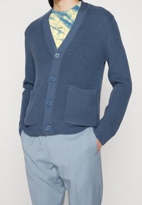 Blåstickad cardigan med V-ringning, fem knappar och två framfickor. Bärs över en tie-dye-t-shirt och matchas med ljusa byxor.