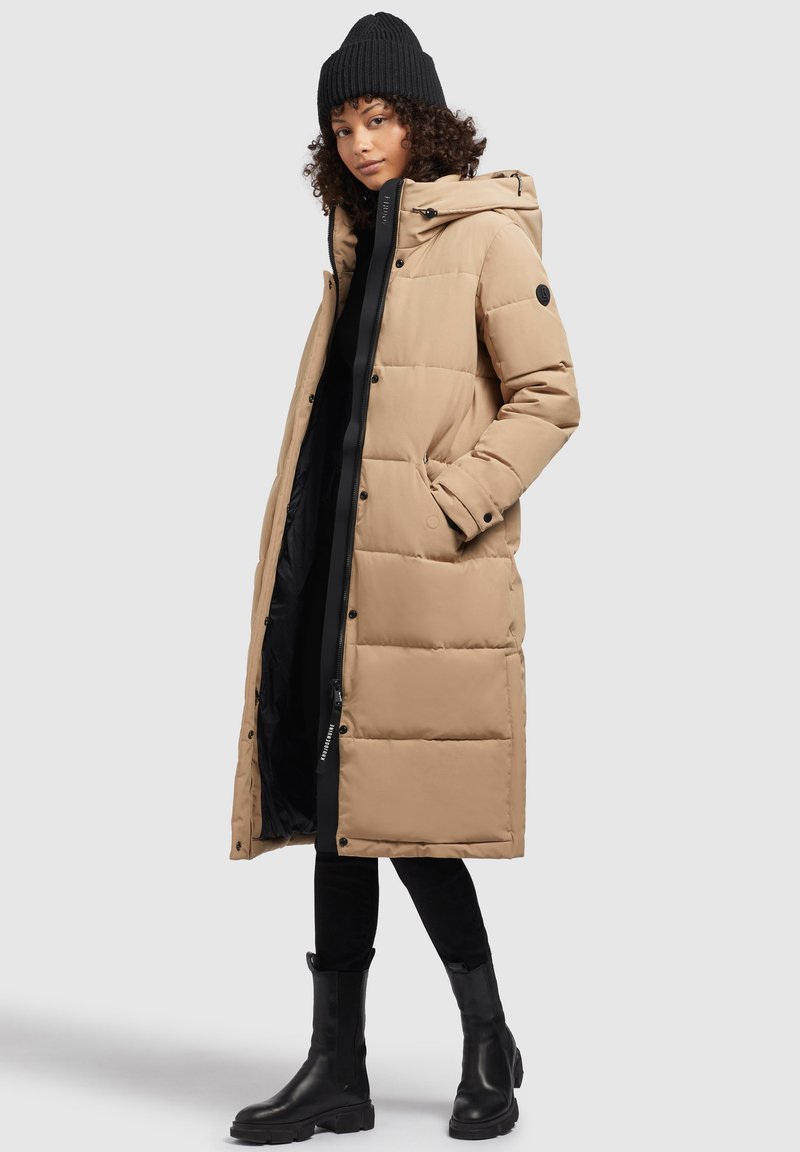 Femme portant un manteau matelassé beige jusqu'aux genoux, un bonnet noir, un pantalon noir et des bottes noires, se tenant avec une main dans la poche de son manteau.