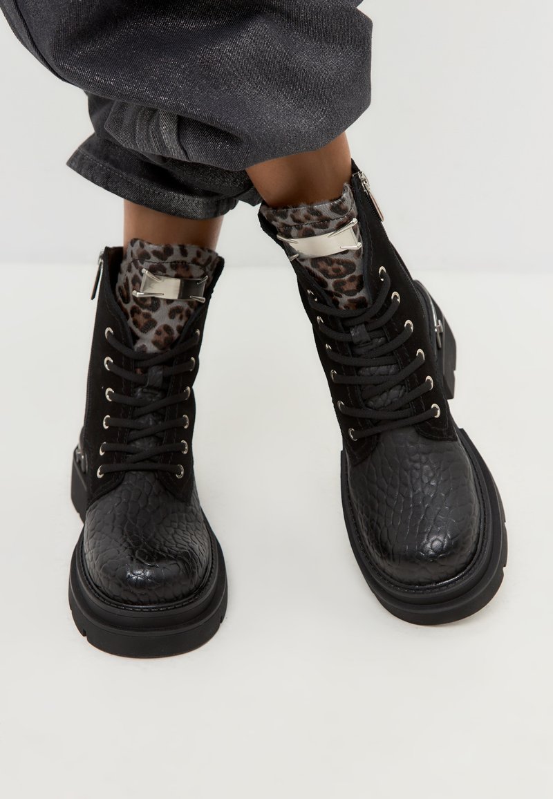 Schwarze Kampfstiefel mit einer strukturierten Lederoberfläche, silbernen Hardware-Akzenten und einem Futter aus Leopardenmuster am oberen Rand.