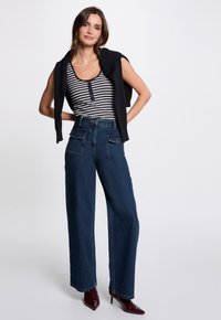 Stribet tanktop med knaplukning, mørkeblå brede bukser med forlommer og burgunder spidse hæle. Modellen står med hænderne på hoften.
