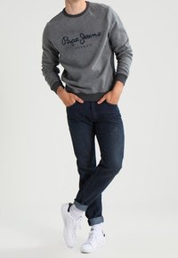 Grauer Sweatshirt mit dunklem Kragen und "Pepe Jeans"-Logo, kombiniert mit dunkelblauen Jeans und weißen Sneakers, hochgekrempelte Bündchen und minimalistisches Design.