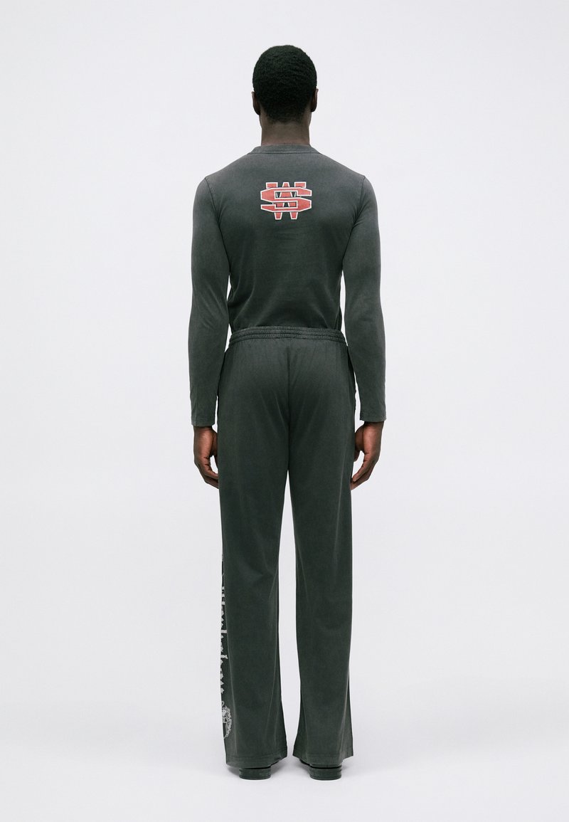 Man som står vänd bort, iklädd en svart långärmad tröja med röd och vit logotyp på den övre delen av ryggen och lösa svarta byxor med sidotryck.