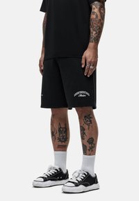Zwarte katoenen shorts met witte logo-tekst, gedragen met een zwart shirt en zwart-witte sneakers. Zichtbare tatoeages op de benen.