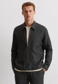Black linen zip-up jacket with a collared neckline, front pocket, and relaxed fit, layered over a light beige shirt. Smooth texture.