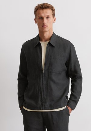 Schwarze Leinen-Zip-Jacke mit Kragen, Fronttasche und lockerer Passform, über einem hellbeigen Shirt getragen. Glatte Textur.