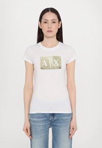 Bílý bavlněný tričko s rectangulárním logem pokrytým zlatými flitry, na kterém jsou bílé písmena "A" a "X." Klasický střih s krátkým rukávem.