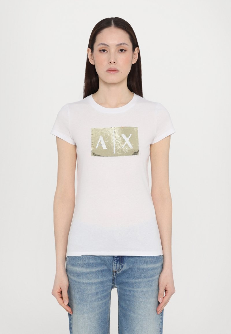 Bílý bavlněný tričko s rectangulárním logem pokrytým zlatými flitry, na kterém jsou bílé písmena "A" a "X." Klasický střih s krátkým rukávem.