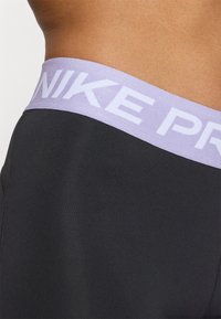 Crne atletske kratke hlače s svijetlopurple elastičnim pojasom na kojem je bijeli tekst "NIKE PRO". Tkanina izgleda glatko i rastezljivo.