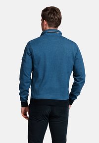 Blauwe gestructureerde ritsjack met een hoge kraag, zwarte afwerking bij de mouwen en taille, en een subtiel ontwerpelement op de achterkant.