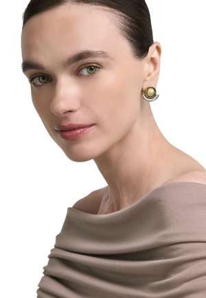 Donna con capelli lisci tirati indietro indossa un top taupe con spalle scoperte e orecchini a mezzo cerchio in oro e argento, guarda dolcemente di lato.