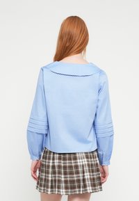 Jasnoniebieska bawełniana bluzka z karbowanym kołnierzykiem i warstwowymi rękawami, zestawiona z brązowo-białą kratkowaną spódnicą.